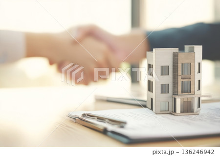 Two hands are shaking over a table that has a contemporary townhouse model and documents. The meeting takes place in an office with natural light coming in from the windows. Two hands are shaking over a table that has a contemporary townhouse model and documents. The meeting takes place in an office with natural light coming in from the windows. 136246942