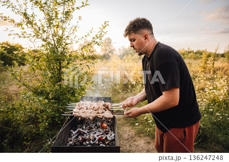 Man grilling pork shashlik over charcoal in countryside at sunset 136247248