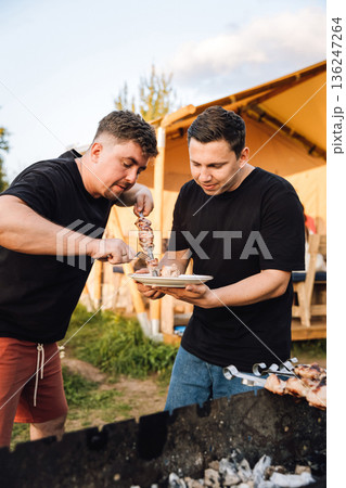 Two friends serving grilled pork shashlik from skewers at an outdoor barbecue 136247264