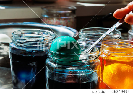 Hand holding spoon with dyed egg taking it out of glass jar during with blue food dye during easter egg coloring 136249495