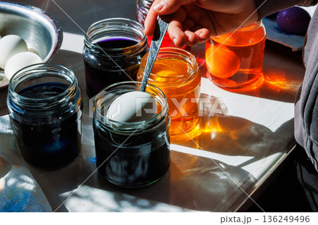 Sunlit close up of a hand using a spoon to dip a white egg into a blue food dye jar, with orange dye jars and other supplies on a kitchen table 136249496