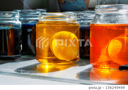 Close up of a easter egg in a yellow and orange food dye solution in a glass jars on a sunlit kitchen table Close up of a easter egg in a yellow and orange food dye solution in a glass jars on a sunlit kitchen table 136249499