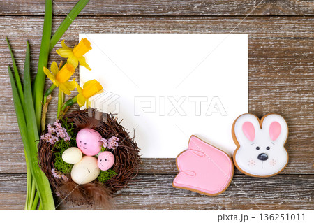 White card on a wooden background with daffodils, nest and Easter cookies. 136251011