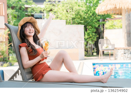プールサイドのリクライニングチェアに座りトロピカルジュースを飲む水着姿の若いアジア人女性 136254902