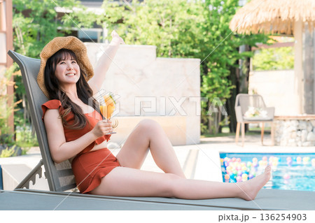 プールサイドのリクライニングチェアに座りトロピカルジュースを飲む水着姿の若いアジア人女性 プールサイドのリクライニングチェアに座りトロピカルジュースを飲む水着姿の若いアジア人女性 136254903