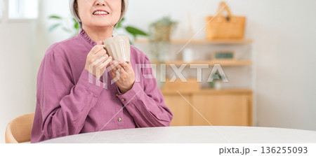 自宅のダイニングテーブルでホットドリンクを飲むシルバーヘアのシニア女性 136255093