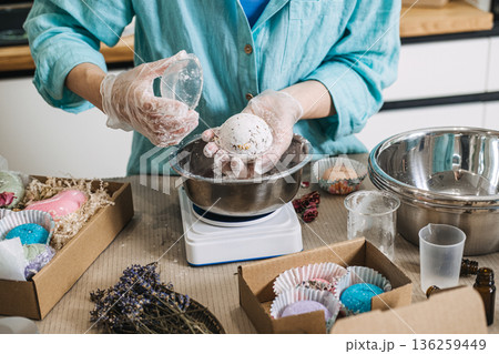 Hands hold large white bath bomb over metal bowl on digital scale surrounded by gift boxes. Small business scale, bulk bath bombs, production day vlog, handmade workshop. Hands hold large white bath bomb over metal bowl on digital scale surrounded by gift boxes. Small business scale, bulk bath bombs, production day vlog, handmade workshop. 136259449