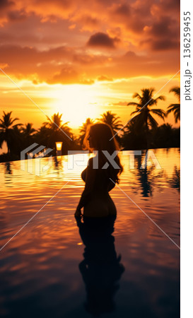 Silhouette of a beautiful woman relaxing in the pool in the setting light of the sun. The pool is surrounded by tropical trees. 136259455