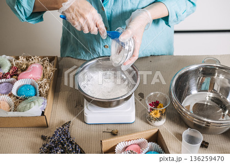 Person pours white powder into stainless steel bowl on digital scale surrounded by colorful finished products. Handmade bath bombs, personalized self-care gifts, bath salt mixology. Person pours white powder into stainless steel bowl on digital scale surrounded by colorful finished products. Handmade bath bombs, personalized self-care gifts, bath salt mixology. 136259470