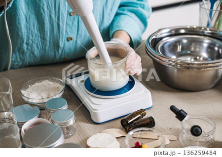 Person using immersion blender to mix liquid base in glass jar on digital scale. cosmetic emulsifying, professional cream blending, skincare laboratory tools, making lotion process. 136259484