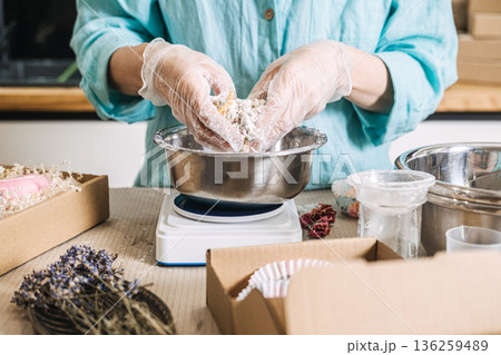 Person mixes botanical ingredients with hands in metal bowl on digital scale surrounded by packaging materials. Clean beauty process, transparent skincare brand, handmade cosmetic workshop 136259489