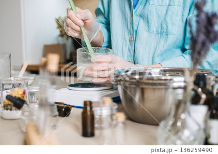 Person in gloves uses spatula to stir liquid in glass container on digital scale. Skincare micro-dosing, active ingredients, precise formulation, botanical potency. 136259490