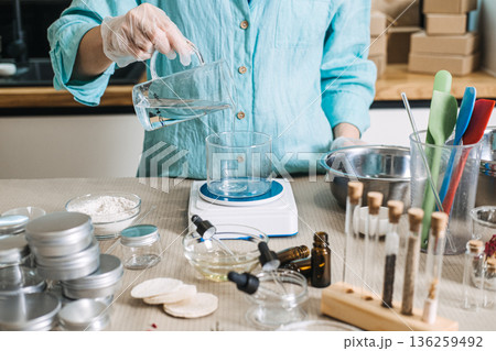 Person pours clear liquid from glass pitcher into glass beaker on digital scale. Cosmetic emulsion physics, oil and water mixing, skincare manufacturing science, professional cream base making. 136259492