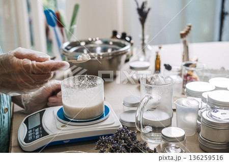 Person in gloves uses wooden spoon to add white solids into glass beaker on digital scale. Artisanal small batch beauty, luxury handmade skincare, exclusive apothecary drops, premium diy cosmetic. Person in gloves uses wooden spoon to add white solids into glass beaker on digital scale. Artisanal small batch beauty, luxury handmade skincare, exclusive apothecary drops, premium diy cosmetic. 136259516