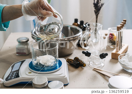 Hand in glove pours liquid oil from small bowl into jar containing white solid butter on digital scale. raw botanical oils, cold pressed skincare, nutrient dense beauty, unrefined ingredients. Hand in glove pours liquid oil from small bowl into jar containing white solid butter on digital scale. raw botanical oils, cold pressed skincare, nutrient dense beauty, unrefined ingredients. 136259519