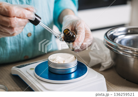 Person uses glass dropper to add liquid into aluminum jar of white cream on digital scale. Clean beauty process, transparent skincare brand, handmade cosmetic workshop, production behind the scenes. Person uses glass dropper to add liquid into aluminum jar of white cream on digital scale. Clean beauty process, transparent skincare brand, handmade cosmetic workshop, production behind the scenes. 136259521
