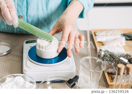 Hands use green spatula to mix white cream in jar on digital scale. Natural skincare formulation, chemical-free cosmetics, botanical ingredients, non-toxic beauty. 136259526