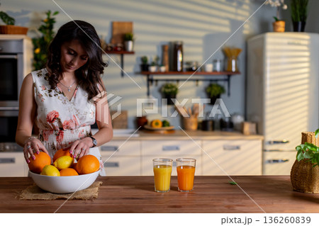 Refreshing morning beverage, Bright kitchen scene with woman pouring fresh orange juice, Morning ambiance with woman making citrus juice amid sunlight and herbal backdrop Refreshing morning beverage, Bright kitchen scene with woman pouring fresh orange juice, Morning ambiance with woman making citrus juice amid sunlight and herbal backdrop 136260839