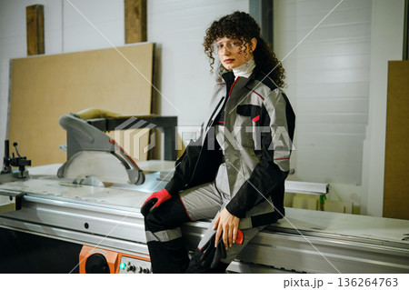 Woman in work outfit sits on a machine in a workshop during the day while wearing gloves 136264763