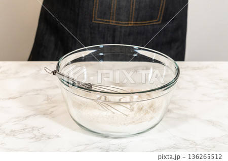Glass mixing bowl containing flour with a metal whisk resting inside. The marble surface and soft lighting create a clean and calm baking preparation scene. Glass mixing bowl containing flour with a metal whisk resting inside. The marble surface and soft lighting create a clean and calm baking preparation scene. 136265512