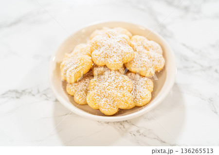 A small white bowl filled with freshly baked lemon spritz cookies, dusted with powdered sugar, sits on a marble countertop. These buttery, melt-in-your-mouth cookies have a delicate texture and a A small white bowl filled with freshly baked lemon spritz cookies, dusted with powdered sugar, sits on a marble countertop. These buttery, melt-in-your-mouth cookies have a delicate texture and a 136265513