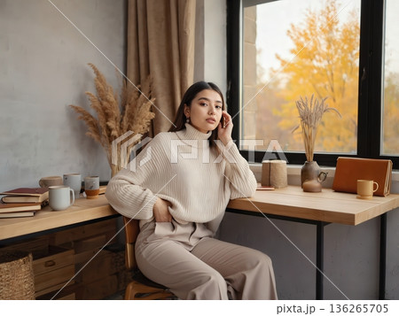 A woman sits at a wooden desk beside a large window showing autumn foliage. Warm natural light fills the room with soft neutral tones. The scene feels cozy, thoughtful, and calm. A woman sits at a wooden desk beside a large window showing autumn foliage. Warm natural light fills the room with soft neutral tones. The scene feels cozy, thoughtful, and calm. 136265705