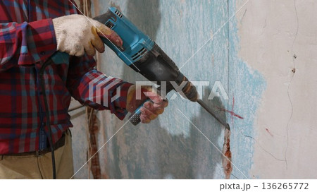 Chipping a wall with a hammer drill in a room undergoing renovation, a worker in brown pants and a checkered shirt holds a jackhammer and chisels a vertical hole in a shabby wall, chiseling a channel Chipping a wall with a hammer drill in a room undergoing renovation, a worker in brown pants and a checkered shirt holds a jackhammer and chisels a vertical hole in a shabby wall, chiseling a channel 136265772