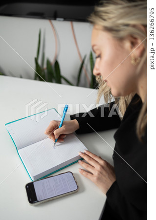 Business Woman Writes in Diary While Sitting at a Desk in an Office Setting Near Plants 136266795