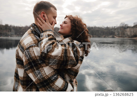 Couple smiles at each other by the lake while wearing matching sweaters on a cloudy day 136268625