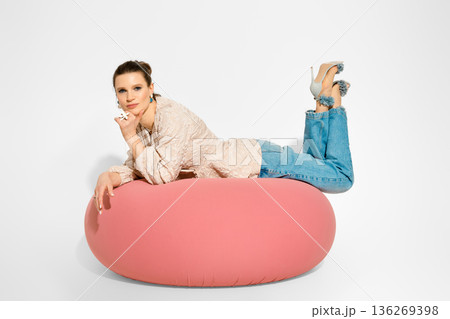 Model poses on pink cushion wearing denim jeans and heels in studio setting Model poses on pink cushion wearing denim jeans and heels in studio setting 136269398