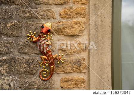 A bright metal lizard sculpture clings to a warm stone wall creating a playful contrast of color and texture in natural light. A bright metal lizard sculpture clings to a warm stone wall creating a playful contrast of color and texture in natural light. 136273842