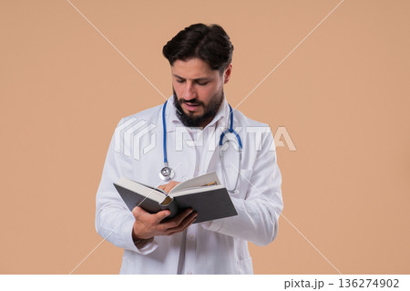 Young Doctor Man Reading Book With Focused Smile On Beige Background. Young Doctor Man Reading Book With Focused Smile On Beige Background. 136274902