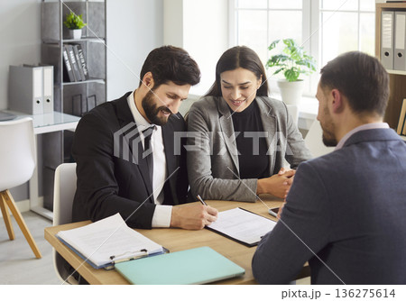 Happy young couple signing contract with male manager while reaching agreement in modern office. Happy young couple signing contract with male manager while reaching agreement in modern office. 136275614