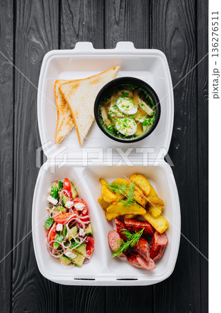 Takeout box with chicken broth soup, toast, fresh salad, potato wedges and sausage 136276511