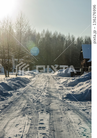 Sunny winter day on a snowy country road with tire tracks and lens flare, leading towards a forest and rural homes. 136276996