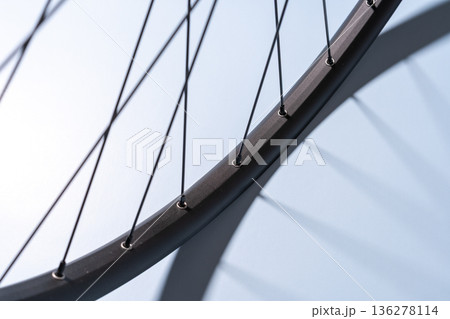 Close-up of a black bicycle rim with metal spokes casting shadows on a light blue surface. 136278114