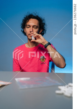 Vertical portrait of young man with curly hair in pink t-shirt using nasal spray to alleviate allergy symptoms or congestion, sitting at a desk snorting drugs from small container with money and pills Vertical portrait of young man with curly hair in pink t-shirt using nasal spray to alleviate allergy symptoms or congestion, sitting at a desk snorting drugs from small container with money and pills 136278882
