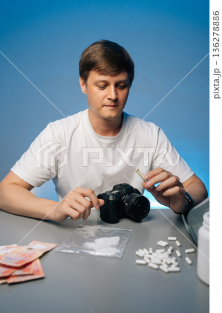 Vertical portrait of photographer preparing drugs with rolled banknote sitting at desk with camera, struggling with addiction, studio shot portrait on isolated background. Concept of substance abuse. Vertical portrait of photographer preparing drugs with rolled banknote sitting at desk with camera, struggling with addiction, studio shot portrait on isolated background. Concept of substance abuse. 136278886