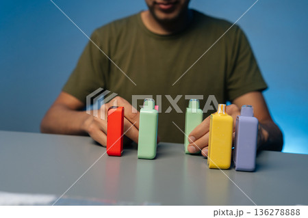 Cropped shot of unrecognizable man holding and presenting various new heat-not-burn tobacco heating systems in vibrant colors on gray table, against isolated blue background. Cropped shot of unrecognizable man holding and presenting various new heat-not-burn tobacco heating systems in vibrant colors on gray table, against isolated blue background. 136278888