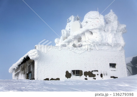The image depicts a remote, utility building with telecommunications antennas and microwave dishes completely buried in snow and ice. 136279398