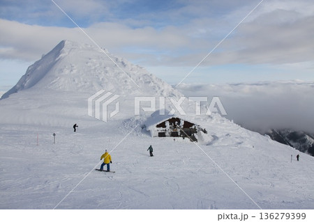 a snow-buried alpine mountain hut on an open ski slope, with snowboarders and skiers passing by 136279399