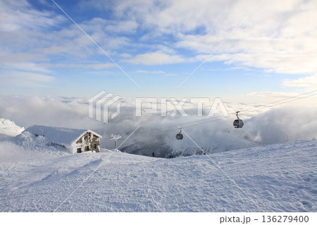 Panoramic winter mountain scene with snow-covered ski resort, cable car gondolas gliding above clouds, alpine hut in foreground, 136279400