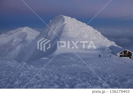 a snow covered mountain peak at dusk, with a dark blue and pink sky and a small mountain hut. 136279403