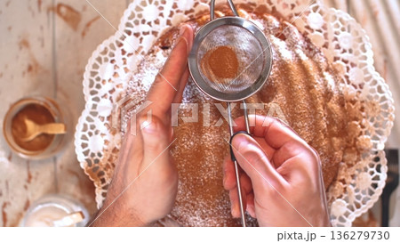 Hands using a strainer to dust powdered sugar on a cake inside a kitchen setting during daylight hours 136279730