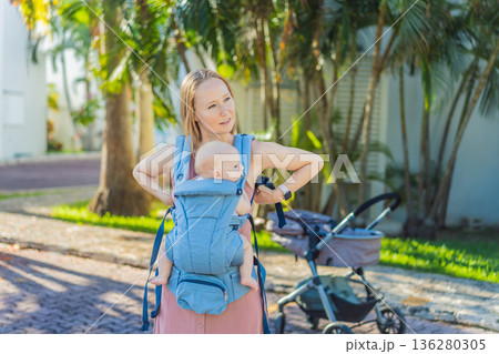 Mother using ergonomic baby carrier during a walk with her infant outdoors. Comfortable babywearing, active motherhood lifestyle, bonding, modern parenting, hands free care and everyday family routine 136280305
