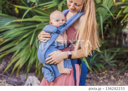 Mother using ergonomic baby carrier during a walk with her infant outdoors. Comfortable babywearing, active motherhood lifestyle, bonding, modern parenting, hands free care and everyday family routine Mother using ergonomic baby carrier during a walk with her infant outdoors. Comfortable babywearing, active motherhood lifestyle, bonding, modern parenting, hands free care and everyday family routine 136282233