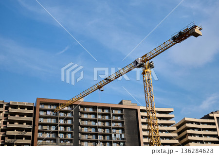 A large yellow tower crane stands over a multi-story residential building under a clear blue sky 136284628