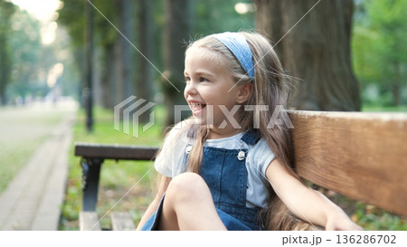 Little happy child girl sitting on a bench smiling happily in summer park. Little happy child girl sitting on a bench smiling happily in summer park. 136286702