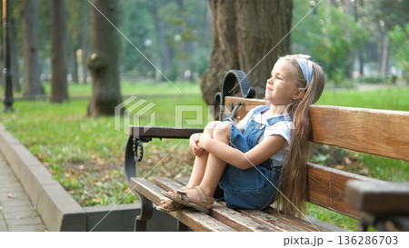 Small happy child girl sitting on a bench resting in summer park. 136286703
