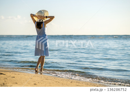 Young woman in straw hat and a dress walking alone on empty sand beach at sea shore. Lonely tourist girl looking at horizon over calm ocean surface on vacation trip. 136286752